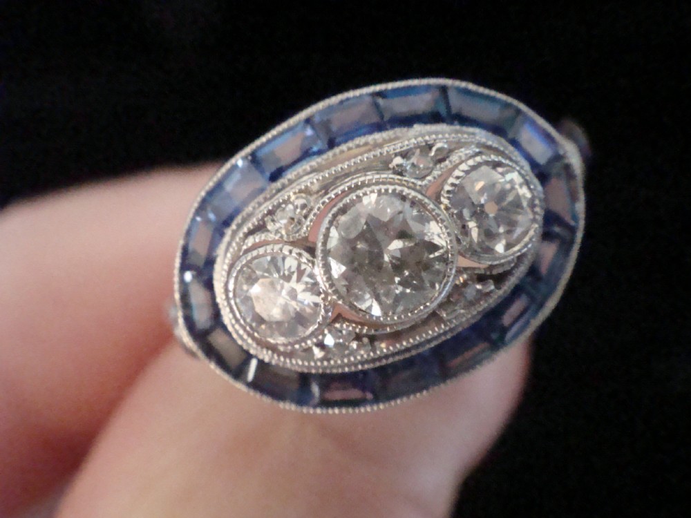 An Art Deco 18ct white gold ring featuring three central diamonds framed by milgrain details and an outer oval border of calibre-cut blue sapphires. View III