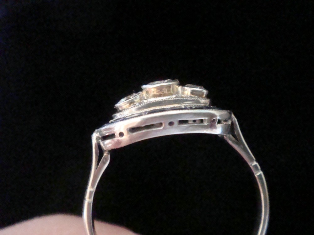 An Art Deco 18ct white gold ring featuring three central diamonds framed by milgrain details and an outer oval border of calibre-cut blue sapphires. View IV