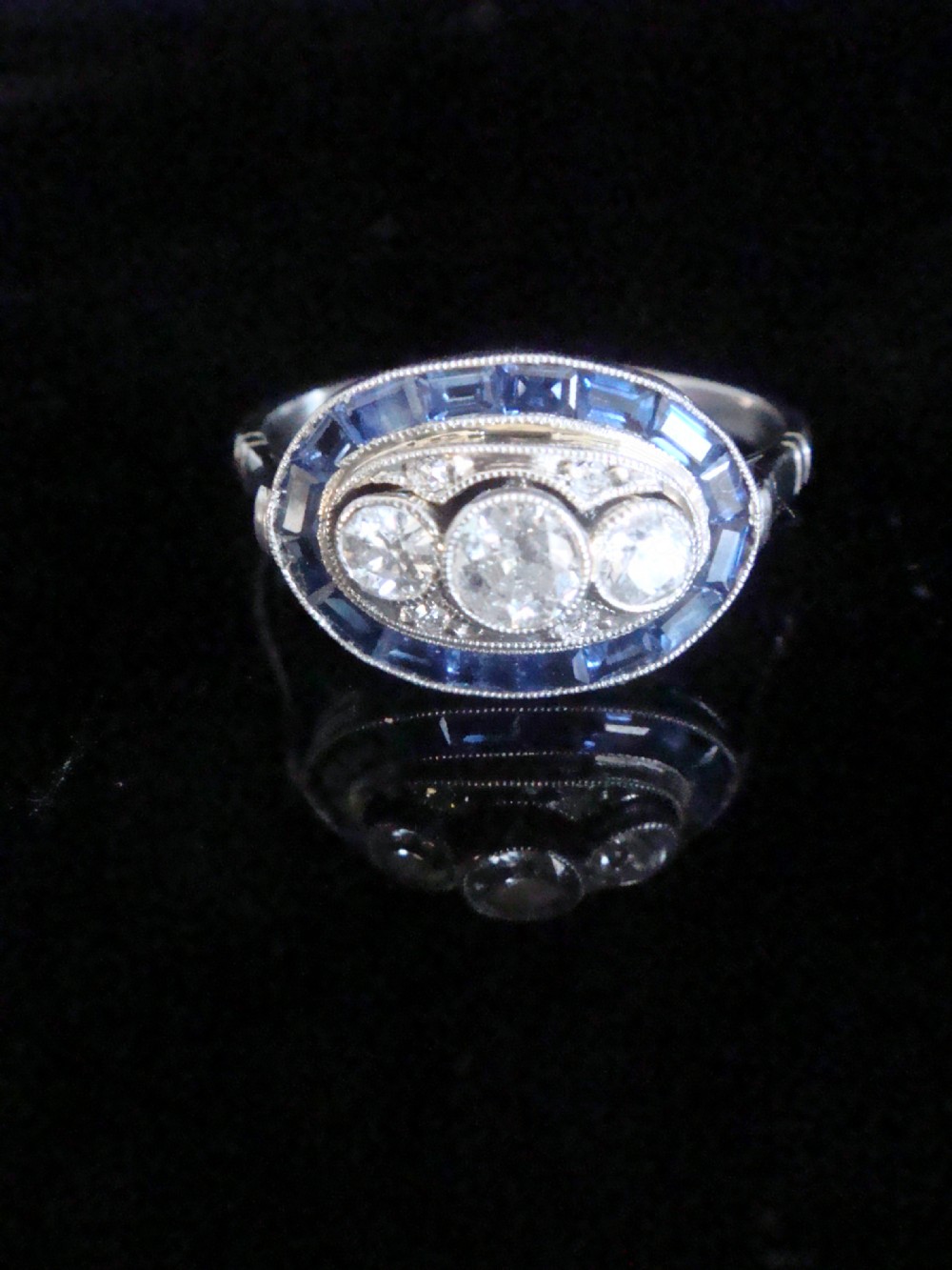 An Art Deco 18ct white gold ring featuring three central diamonds framed by milgrain details and an outer oval border of calibre-cut blue sapphires. View V