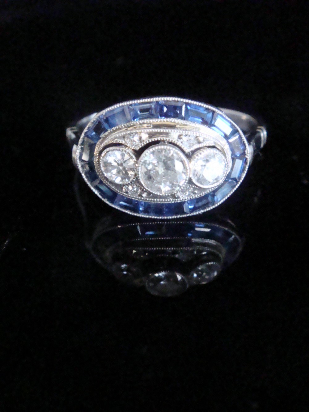 An Art Deco 18ct white gold ring featuring three central diamonds framed by milgrain details and an outer oval border of calibre-cut blue sapphires. View VI