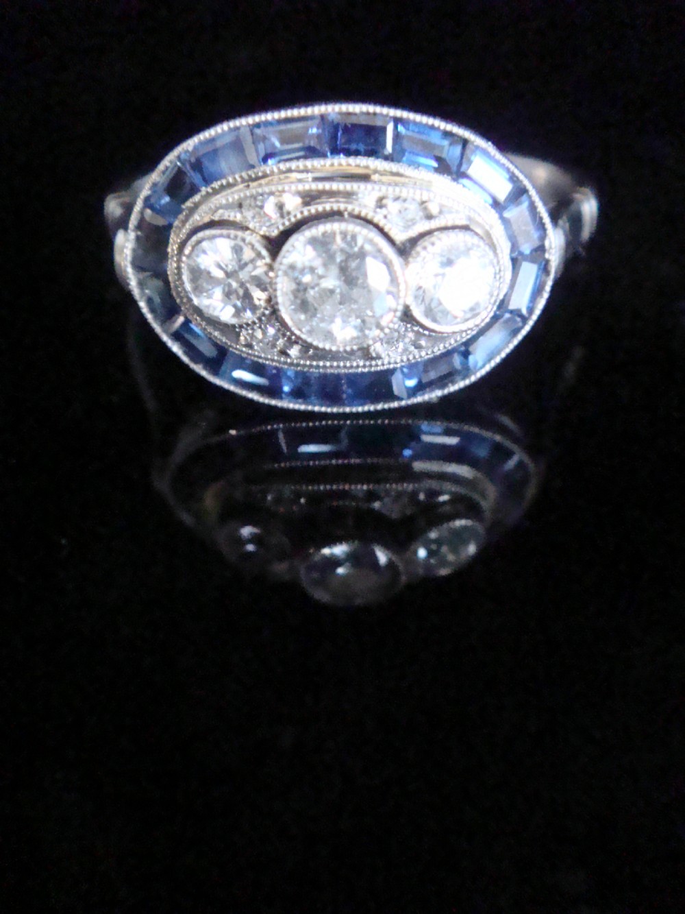 An Art Deco 18ct white gold ring featuring three central diamonds framed by milgrain details and an outer oval border of calibre-cut blue sapphires. View VII