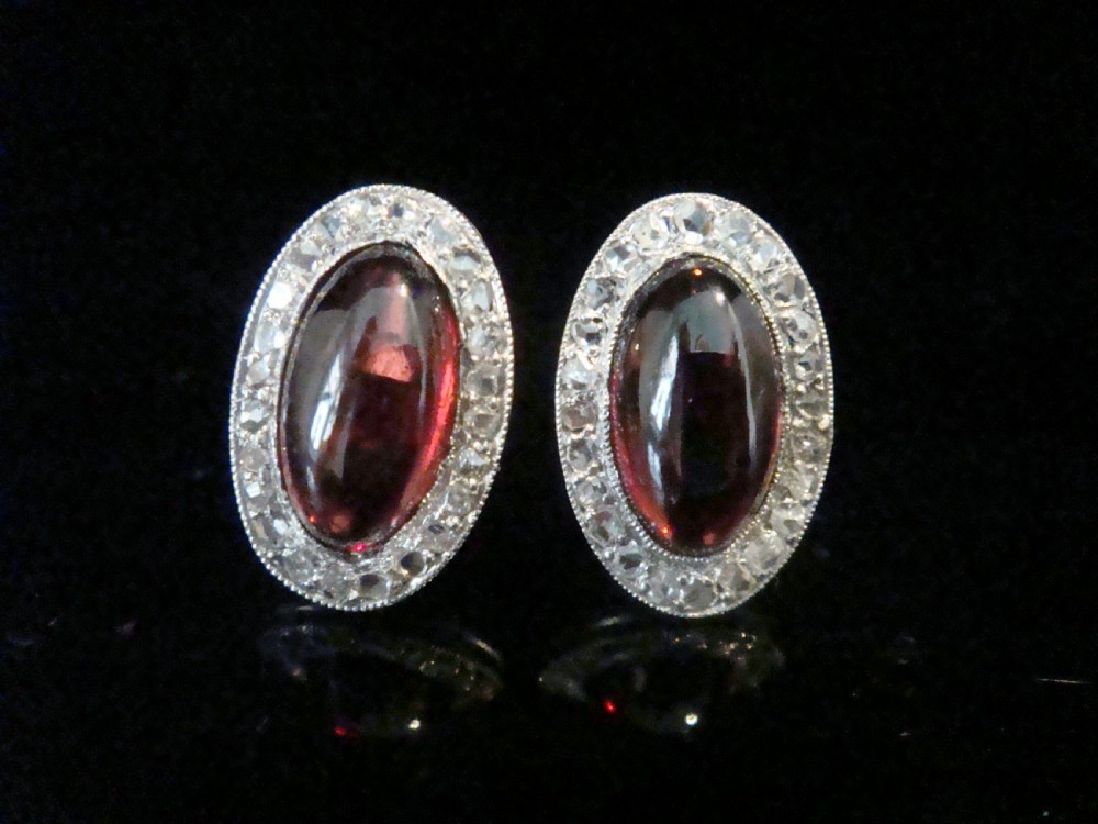 A pair of oval Edwardian stud earrings featuring central smooth cabochon red garnets surrounded by halos of small rose-cut diamonds set in millegrained white metal.