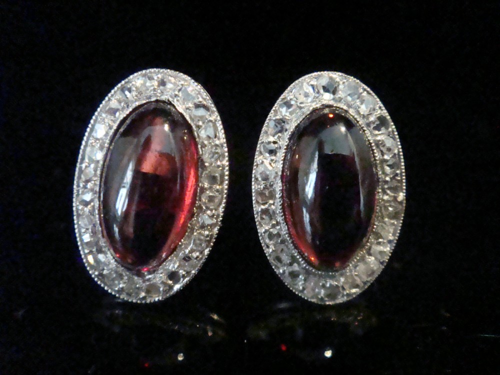 A pair of oval Edwardian stud earrings featuring central smooth cabochon red garnets surrounded by halos of small rose-cut diamonds set in millegrained white metal. View II