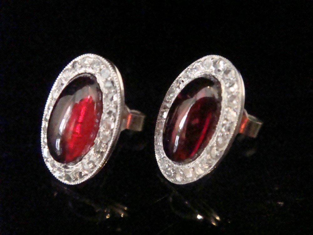 A pair of oval Edwardian stud earrings featuring central smooth cabochon red garnets surrounded by halos of small rose-cut diamonds set in millegrained white metal. View III