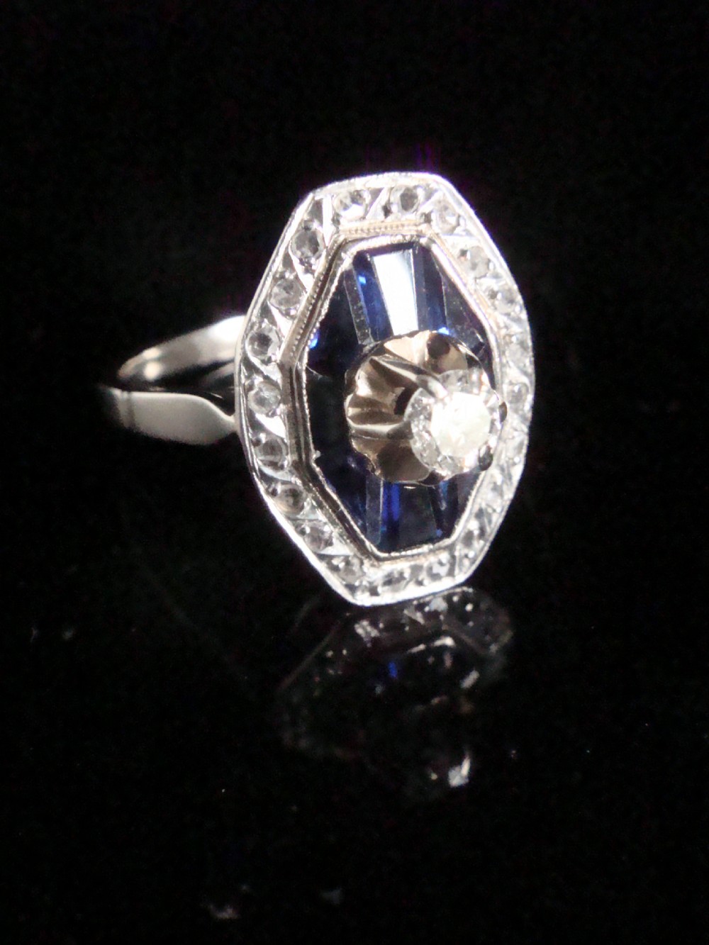 An Art Deco 18ct white gold ring featuring a central diamond encircled by dark blue calibrated sapphires and an outer geometric diamond bezel.