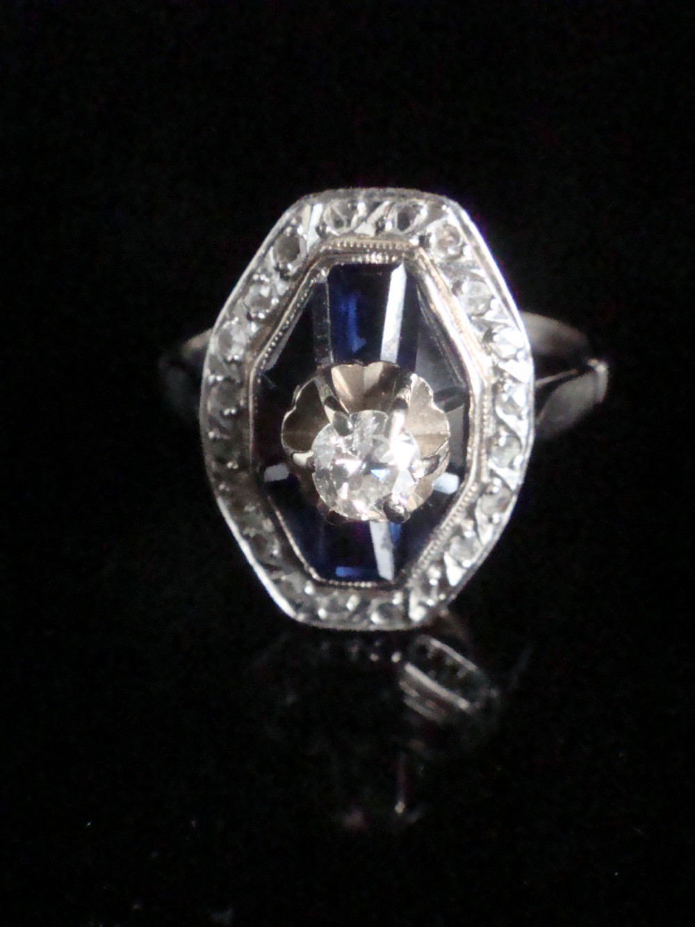 An Art Deco 18ct white gold ring featuring a central diamond encircled by dark blue calibrated sapphires and an outer geometric diamond bezel. View II