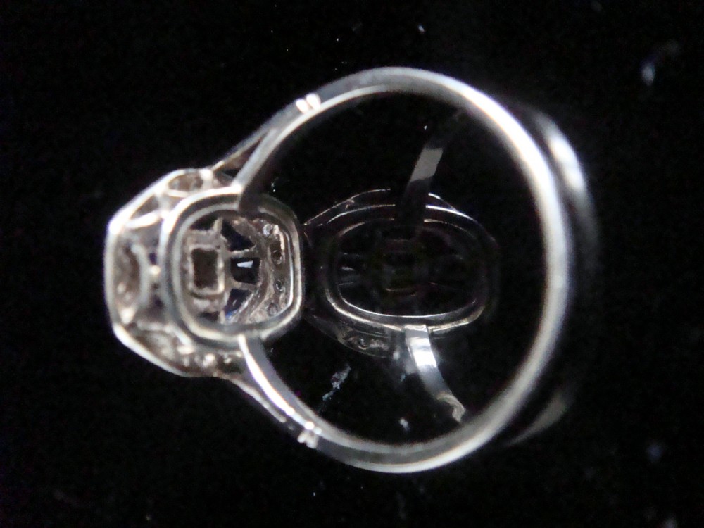 An Art Deco 18ct white gold ring featuring a central diamond encircled by dark blue calibrated sapphires and an outer geometric diamond bezel. View IV