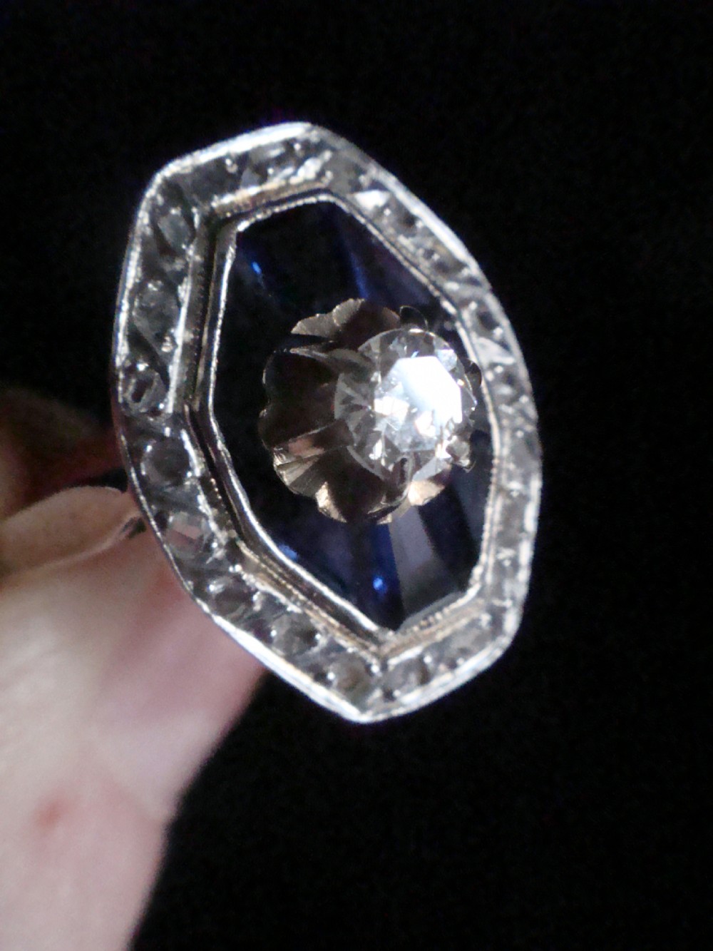 An Art Deco 18ct white gold ring featuring a central diamond encircled by dark blue calibrated sapphires and an outer geometric diamond bezel. View V