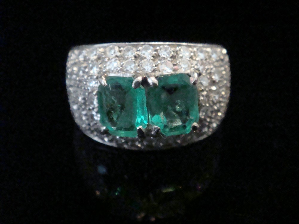 A wide 18ct white gold Retro tank ring featuring two emerald-cut green emeralds set at the centre, surrounded by pave-set round diamonds.