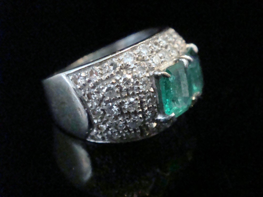 A wide 18ct white gold Retro tank ring featuring two emerald-cut green emeralds set at the centre, surrounded by pave-set round diamonds. View II