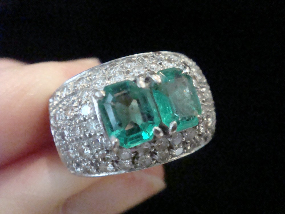 A wide 18ct white gold Retro tank ring featuring two emerald-cut green emeralds set at the centre, surrounded by pave-set round diamonds. View IV