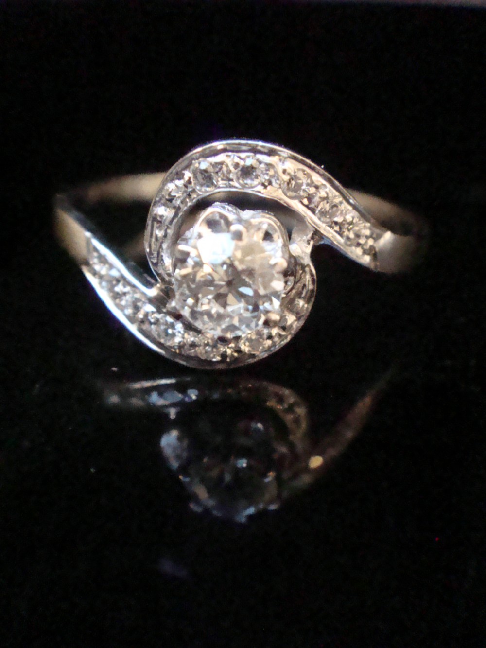 An Edwardian 18ct white gold solitaire ring with a fluid swirl design set with a central old cut diamond and pave accents.
