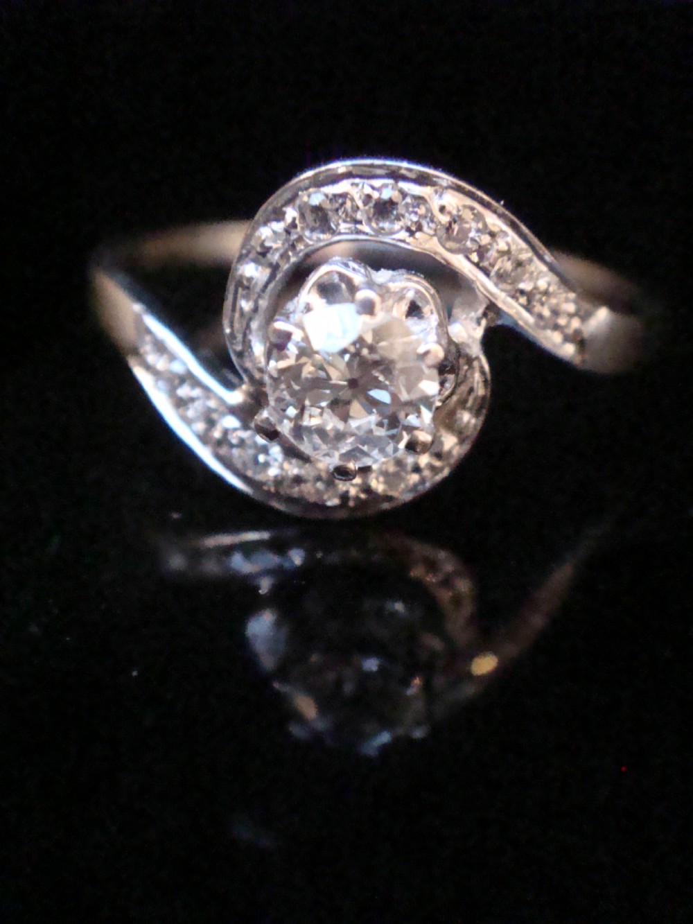 An Edwardian 18ct white gold solitaire ring with a fluid swirl design set with a central old cut diamond and pave accents. View II