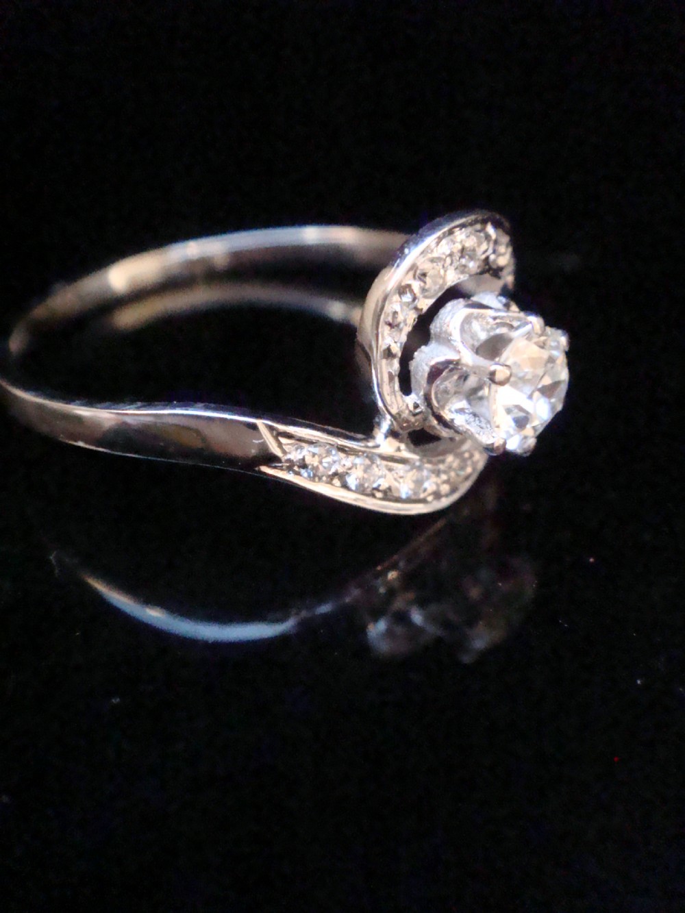 An Edwardian 18ct white gold solitaire ring with a fluid swirl design set with a central old cut diamond and pave accents. View III