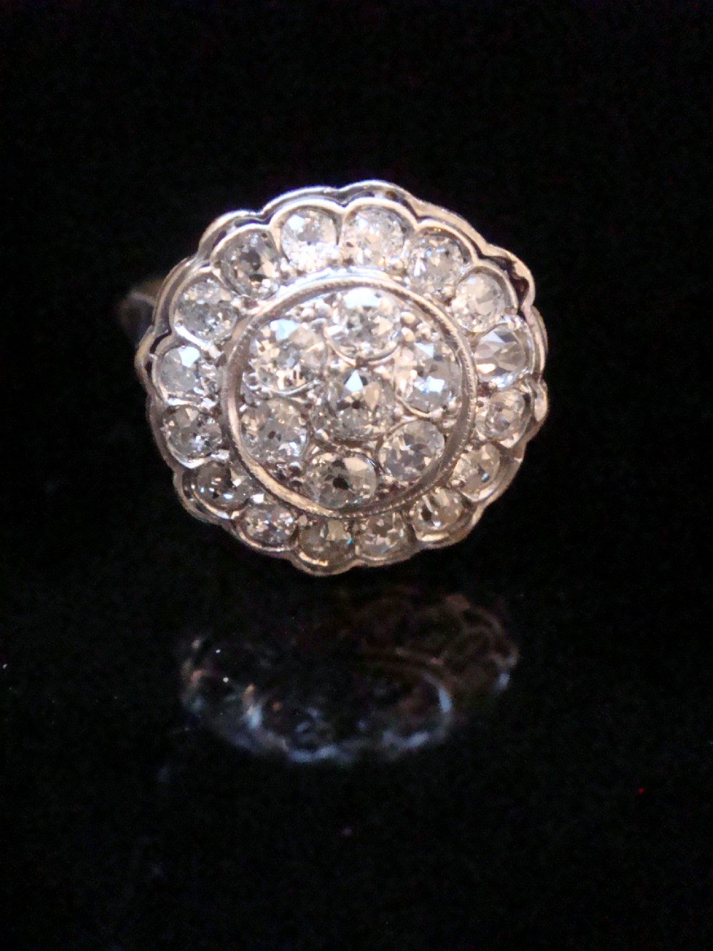 A circular 18ct white gold Edwardian cluster ring set with multiple old cut diamonds featuring a scalloped edge.