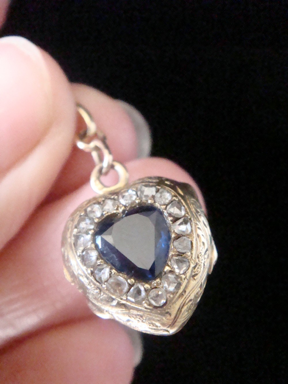 A heart-shaped 18ct gold Edwardian locket set with a central blue sapphire framed by small diamonds and detailed edge engraving.