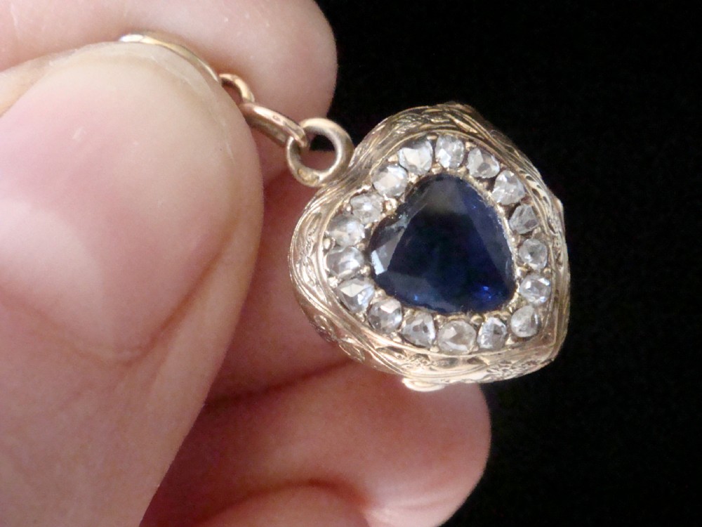 A heart-shaped 18ct gold Edwardian locket set with a central blue sapphire framed by small diamonds and detailed edge engraving. View VI