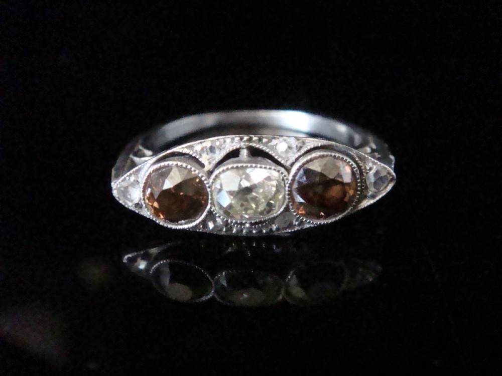 An antique platinum ring featuring a central round white diamond flanked by two natural brown diamonds with millegrain borders.