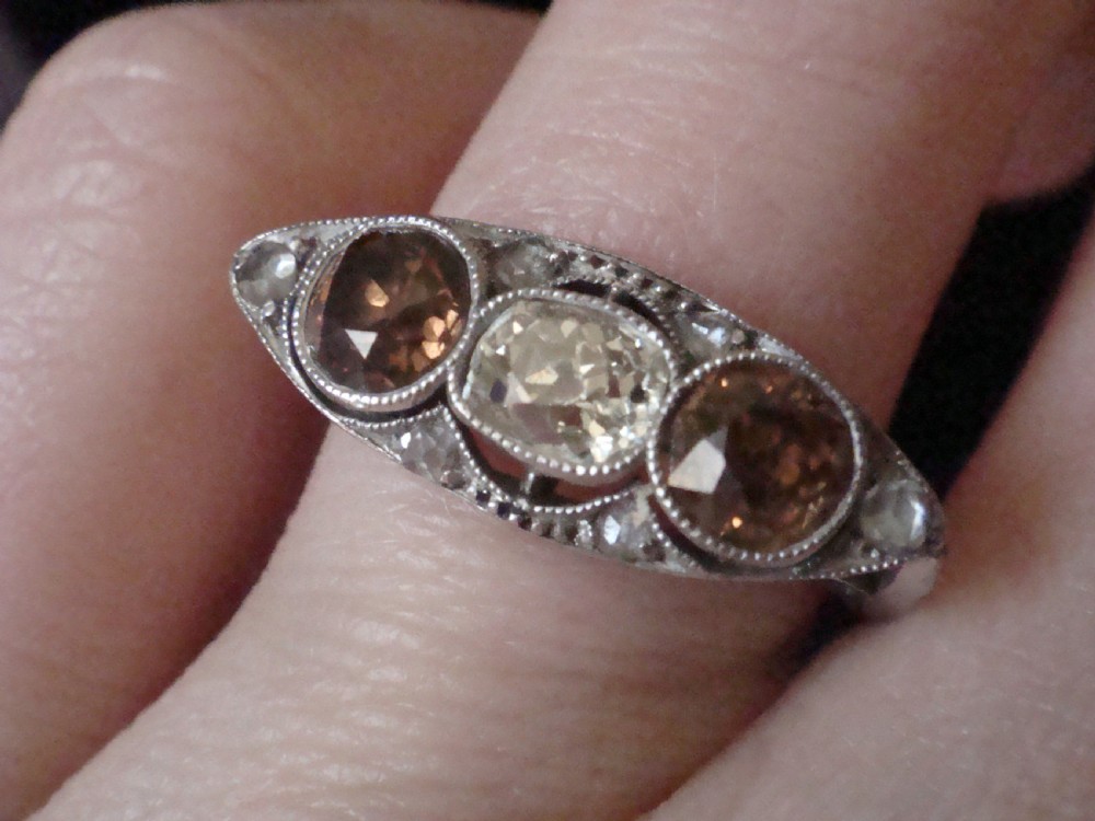 An antique platinum ring featuring a central round white diamond flanked by two natural brown diamonds with millegrain borders. View IV