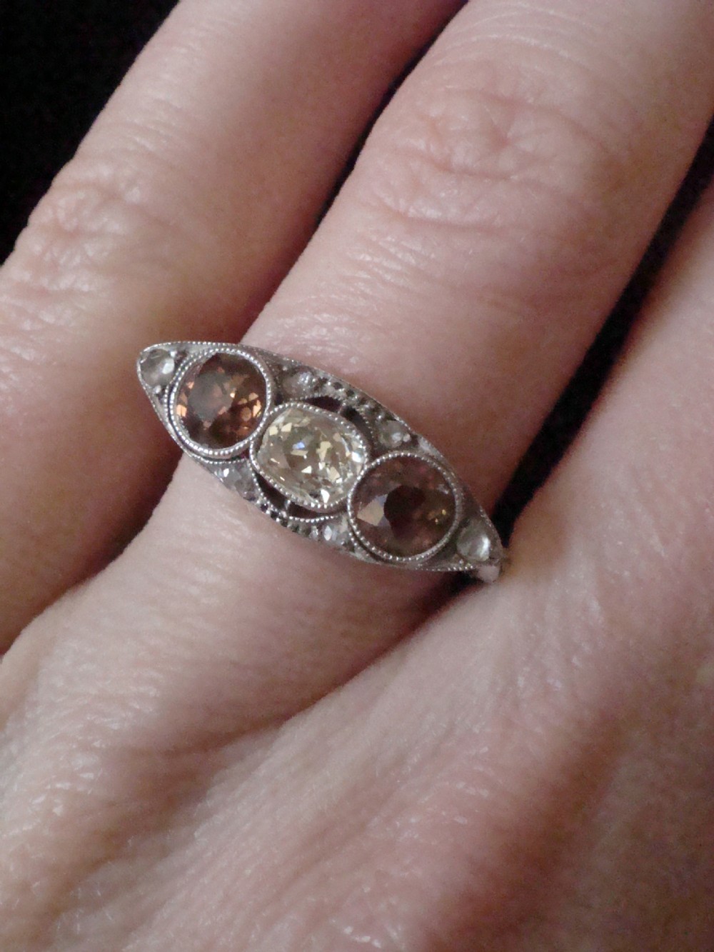 An antique platinum ring featuring a central round white diamond flanked by two natural brown diamonds with millegrain borders. View IX