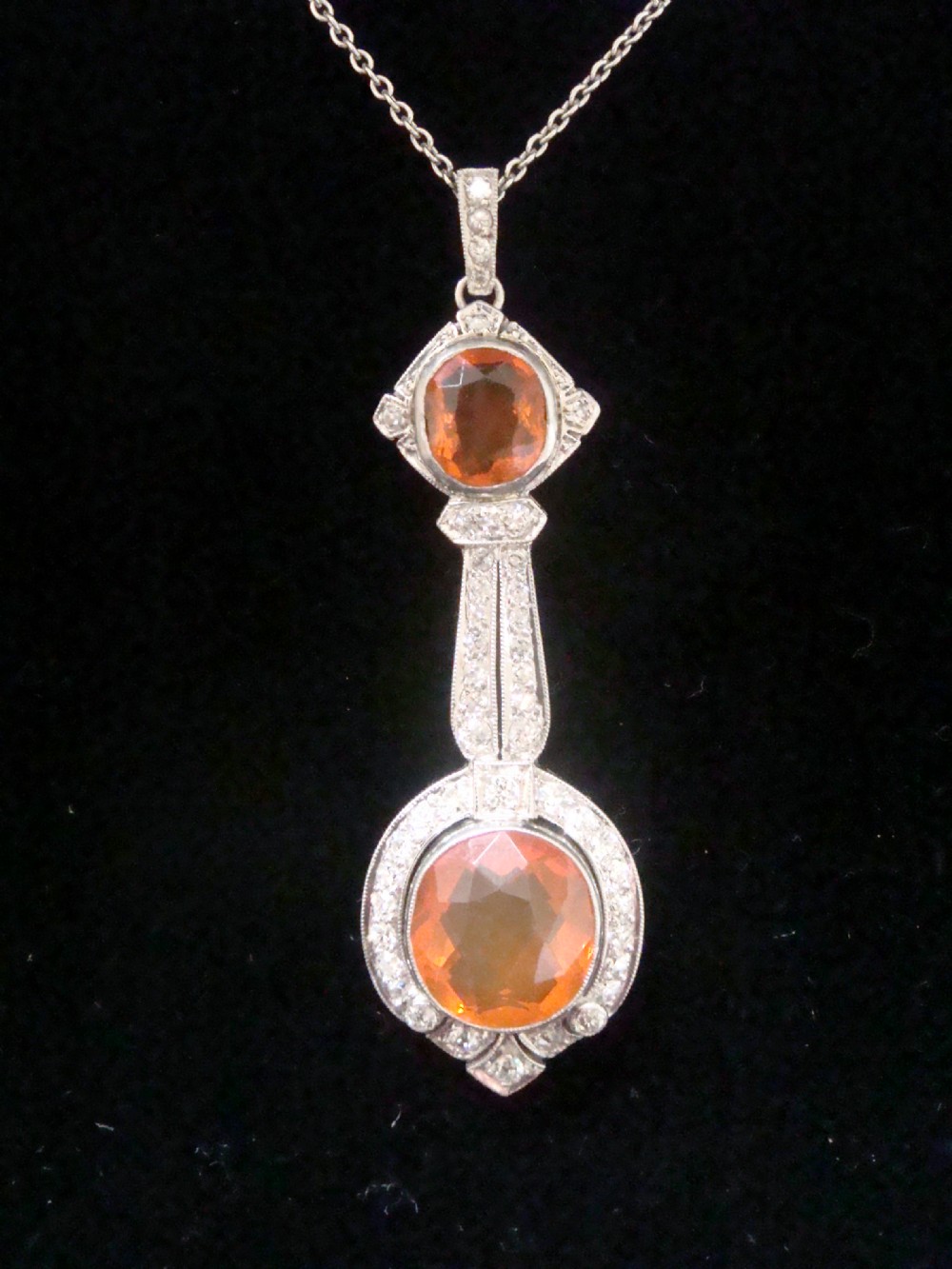 An Edwardian 18ct white gold pendant necklace featuring two oval cut orange fire opals framed by small diamonds along an articulated geometric drop.