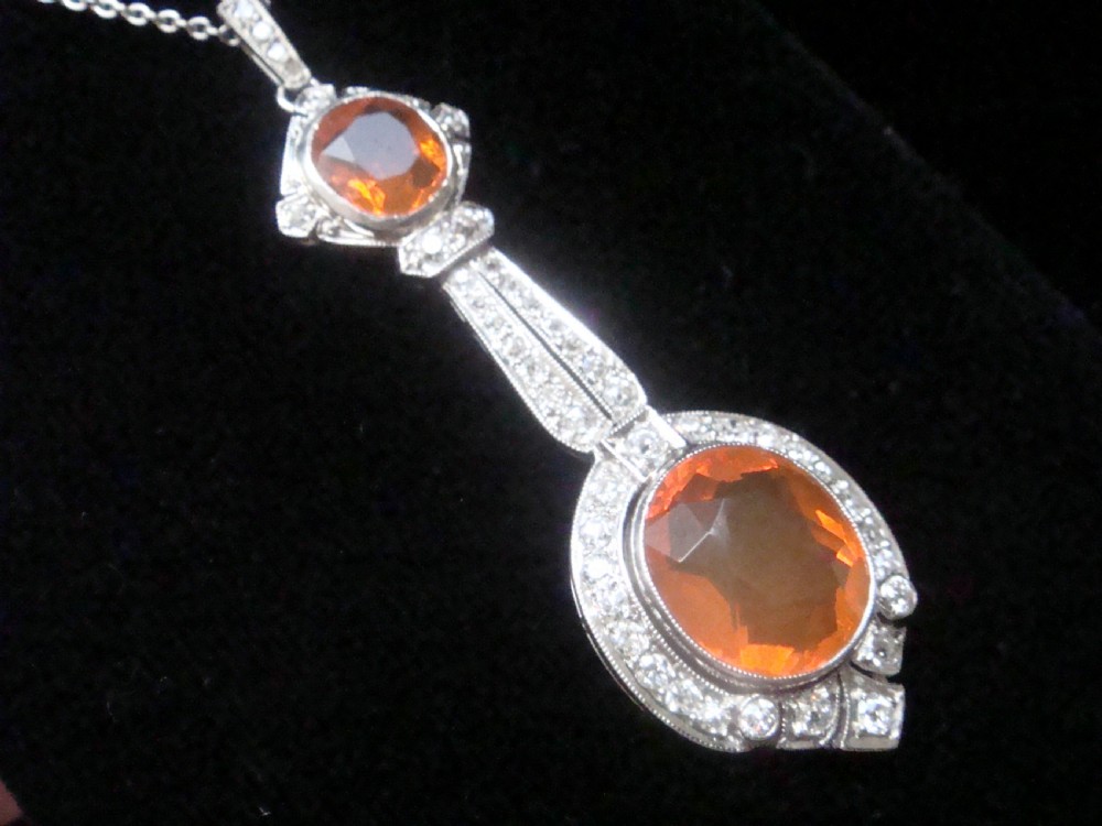 An Edwardian 18ct white gold pendant necklace featuring two oval cut orange fire opals framed by small diamonds along an articulated geometric drop. View V