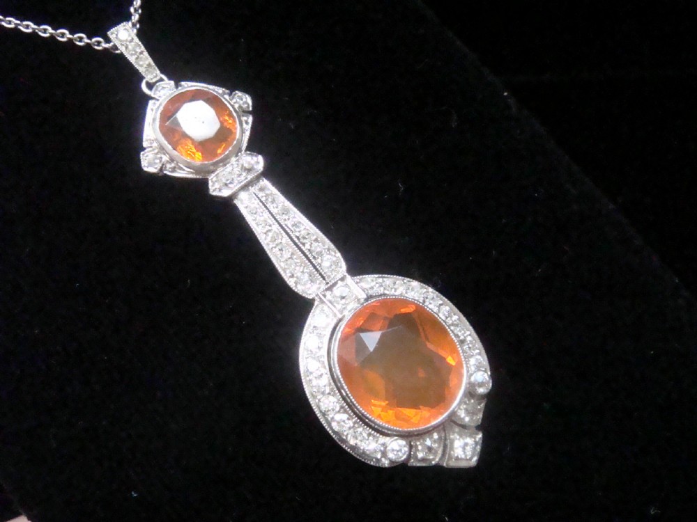 An Edwardian 18ct white gold pendant necklace featuring two oval cut orange fire opals framed by small diamonds along an articulated geometric drop. View VI