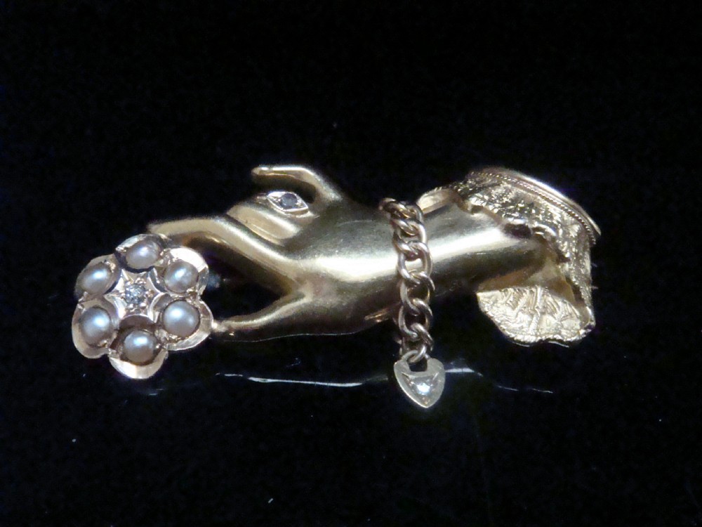 A Victorian 15ct yellow gold brooch carved in the shape of a hand with a lace cuff holding a seed pearl flower with a small heart padlock charm attached.