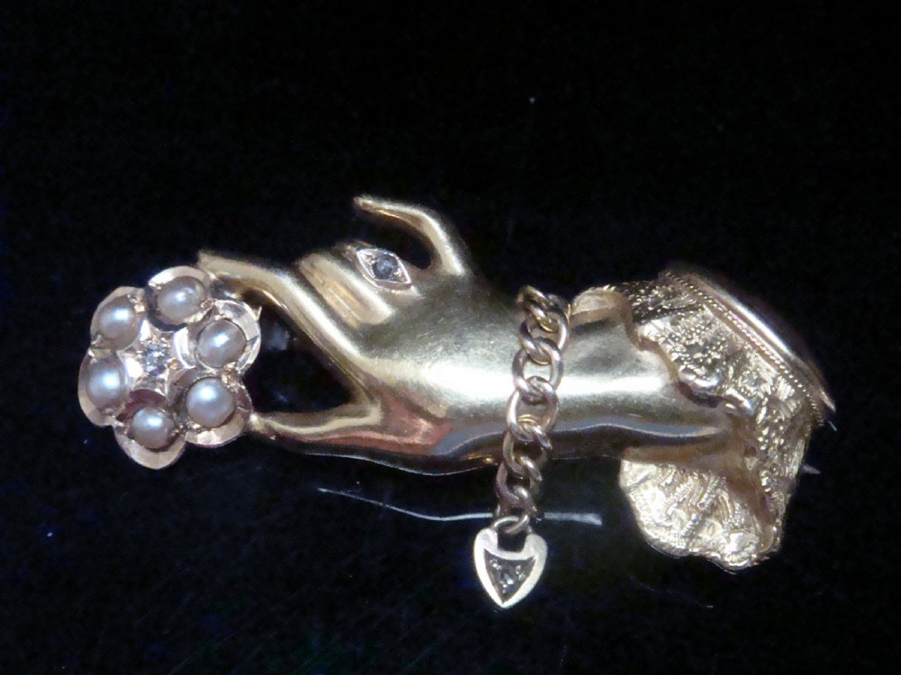 A Victorian 15ct yellow gold brooch carved in the shape of a hand with a lace cuff holding a seed pearl flower with a small heart padlock charm attached. View IV