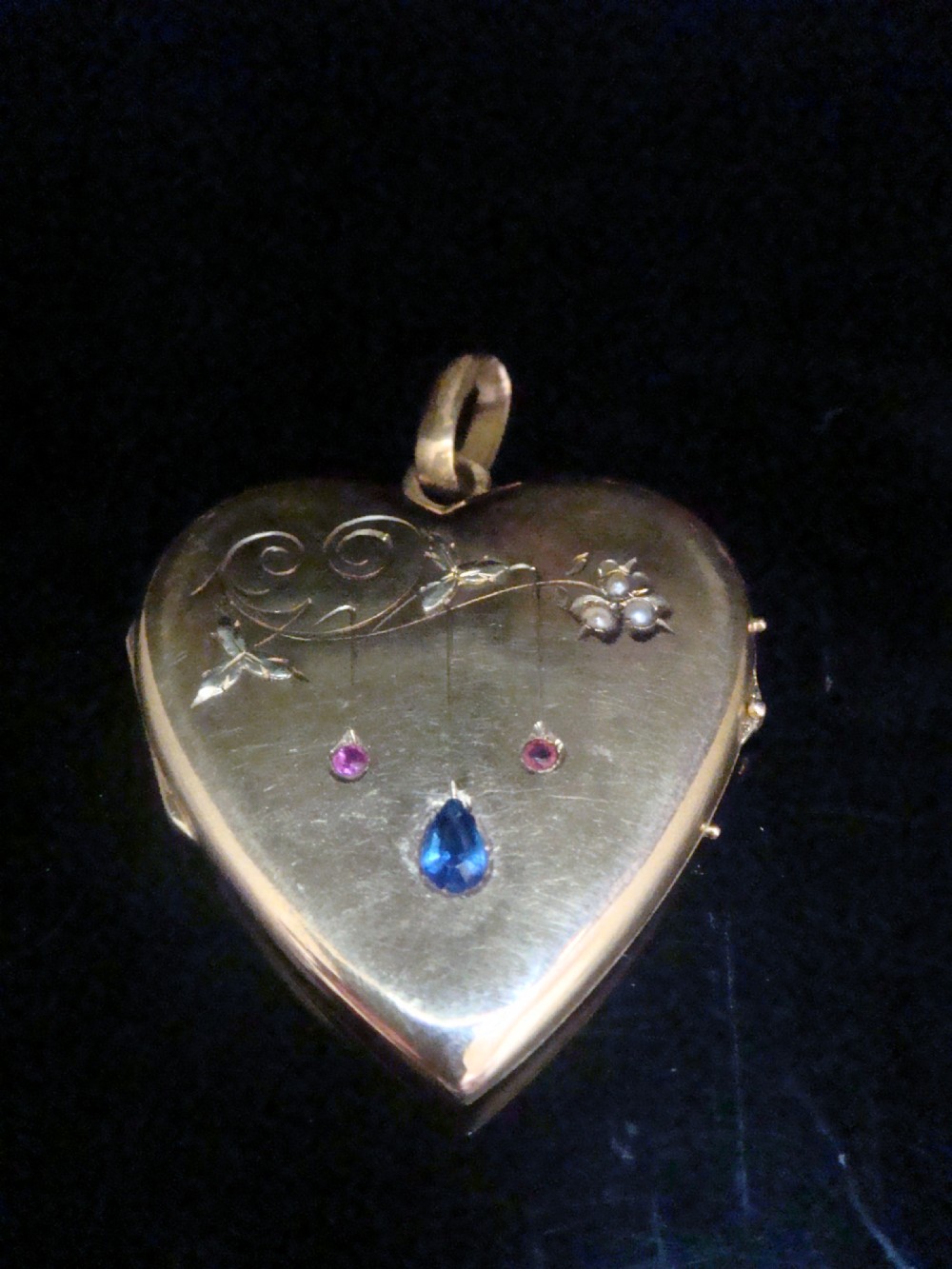 A heart-shaped 18ct yellow gold locket decorated with botanical engravings, a central pear-cut blue sapphire, two small rubies, and seed pearls.