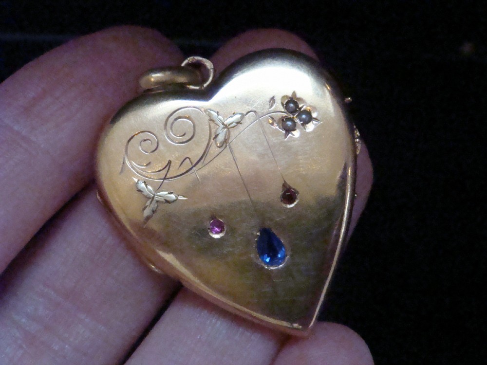 A heart-shaped 18ct yellow gold locket decorated with botanical engravings, a central pear-cut blue sapphire, two small rubies, and seed pearls. View VI