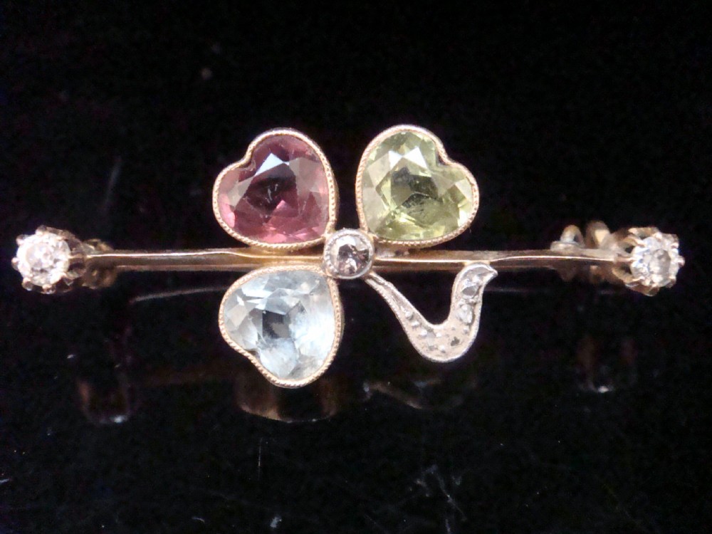 A Victorian 15ct yellow gold bar brooch featuring a central three-leaf clover crafted from heart-shaped tourmaline, aquamarine, and peridot, detailed with diamonds.