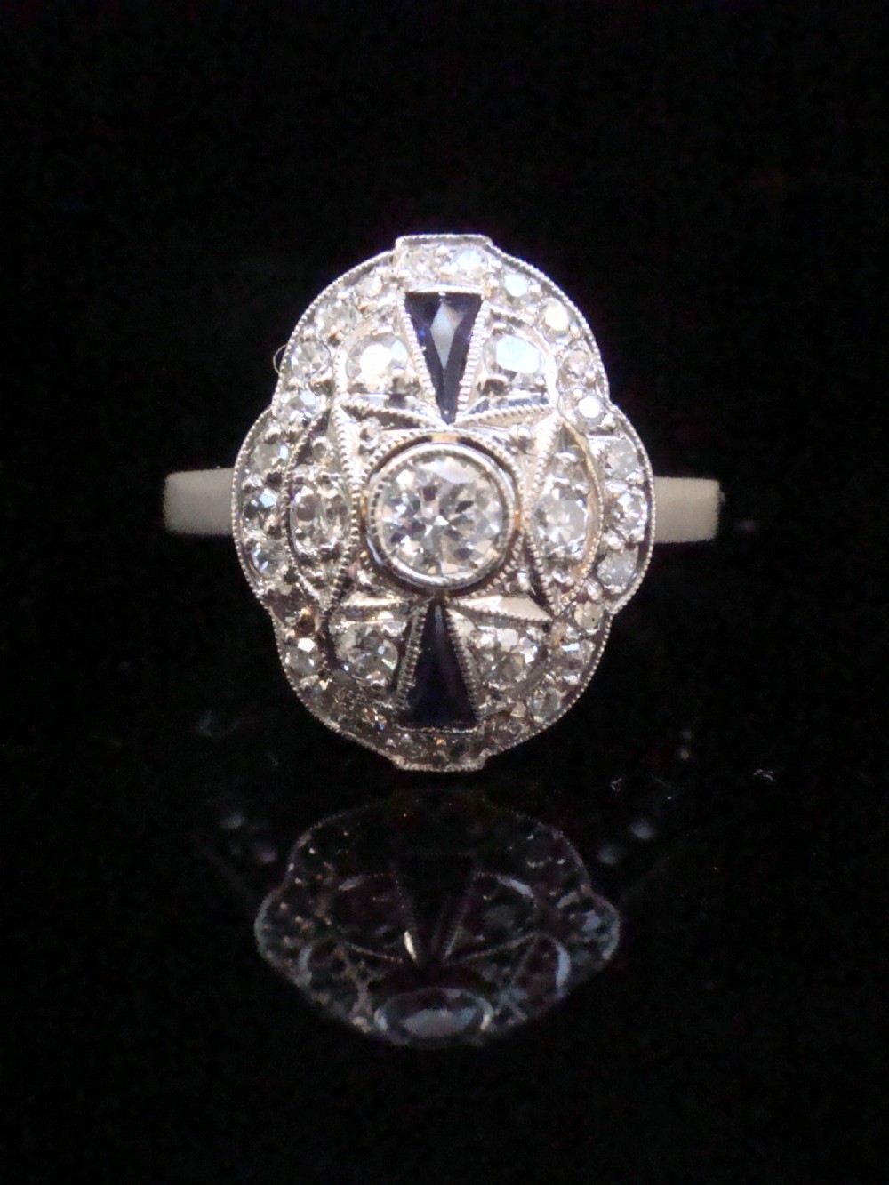 An Art Deco 18ct white gold oval cluster ring featuring central diamonds framed by geometric calibre-cut blue sapphires.