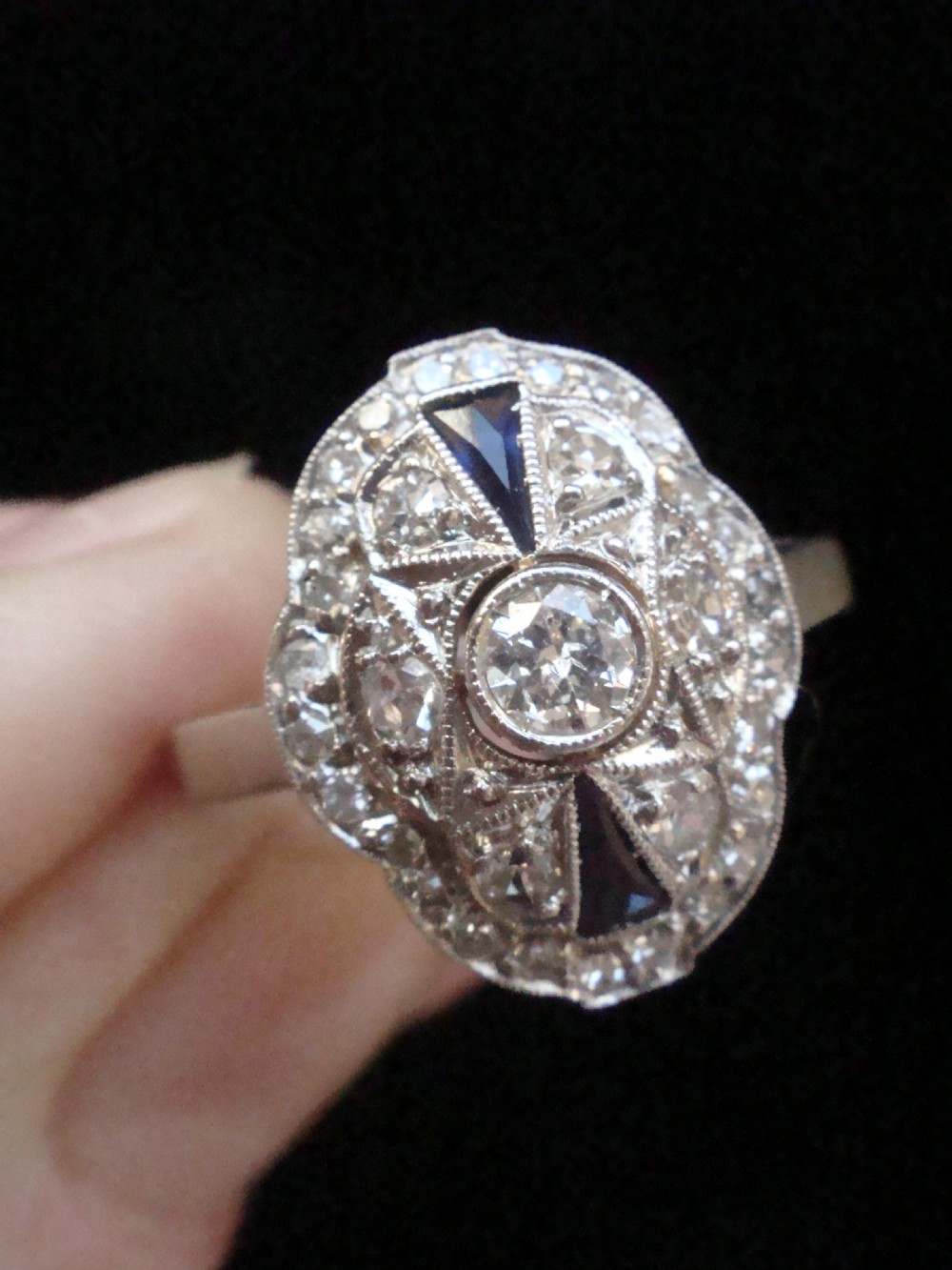 An Art Deco 18ct white gold oval cluster ring featuring central diamonds framed by geometric calibre-cut blue sapphires. View III