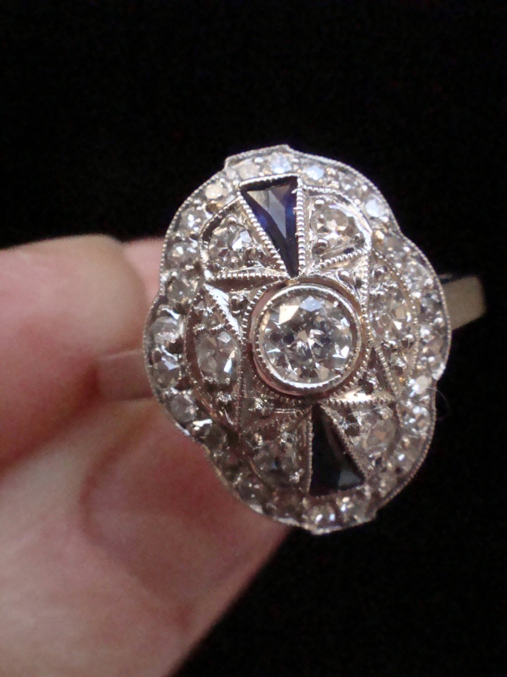 An Art Deco 18ct white gold oval cluster ring featuring central diamonds framed by geometric calibre-cut blue sapphires. View IV