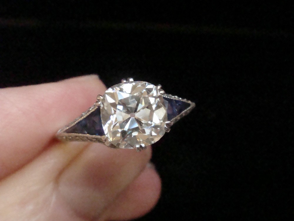 An Art Deco platinum solitaire ring featuring a large transitional-cut central diamond flanked by two triangular blue sapphires.