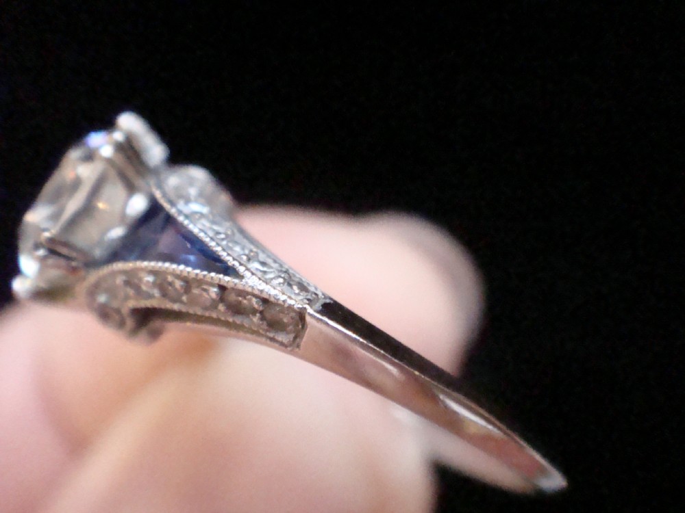 An Art Deco platinum solitaire ring featuring a large transitional-cut central diamond flanked by two triangular blue sapphires. View IV