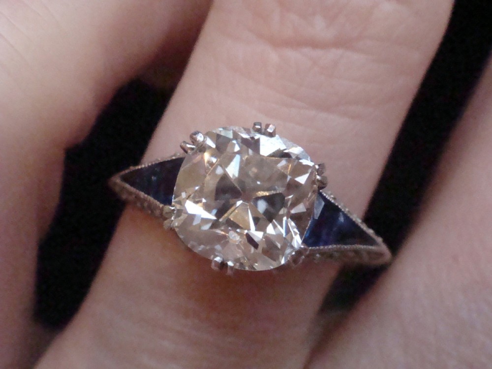 An Art Deco platinum solitaire ring featuring a large transitional-cut central diamond flanked by two triangular blue sapphires. View X