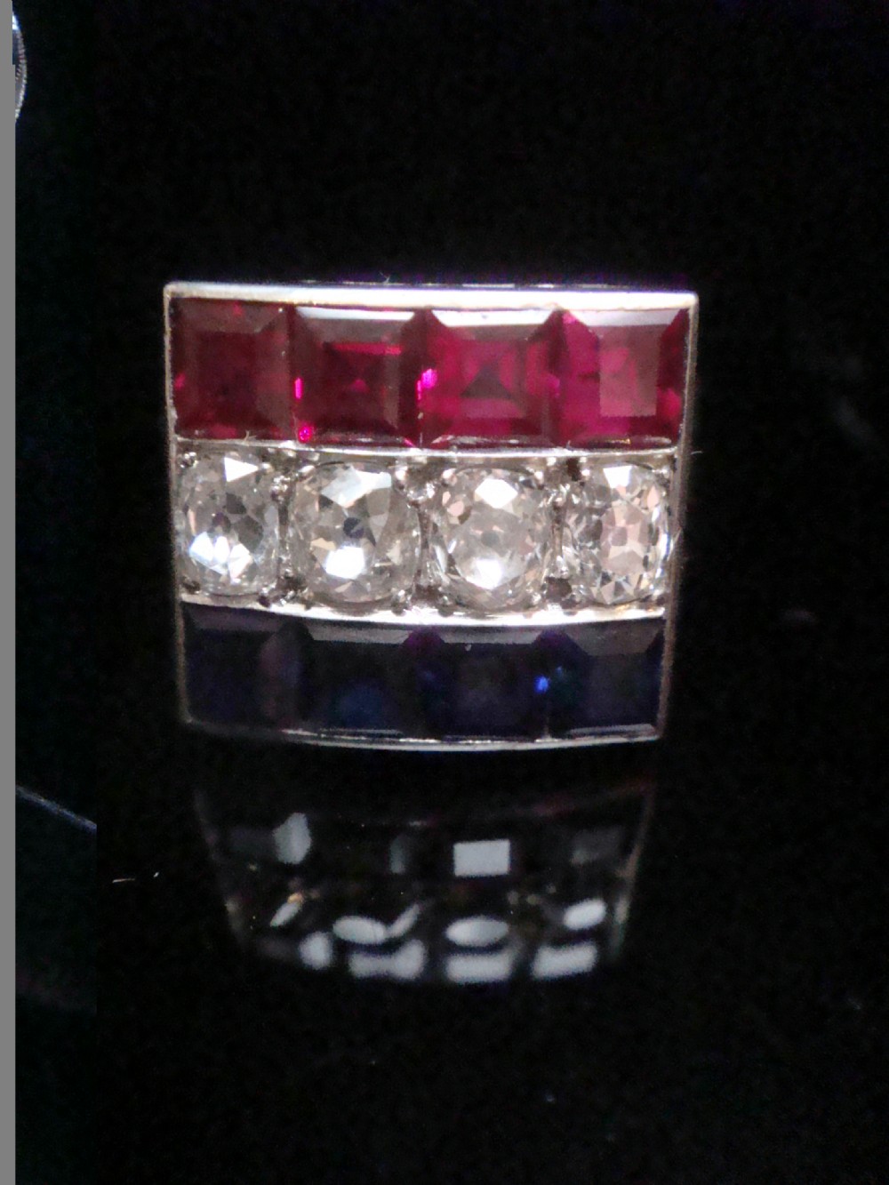An Art Deco platinum rectangular plaque ring set with horizontal rows of red rubies, white diamonds, and blue sapphires.