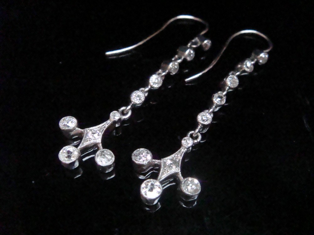 Pair of Edwardian platinum drop earrings featuring articulated lines of bezel-set round old cut diamonds terminating in geometric drops with hook fastenings.