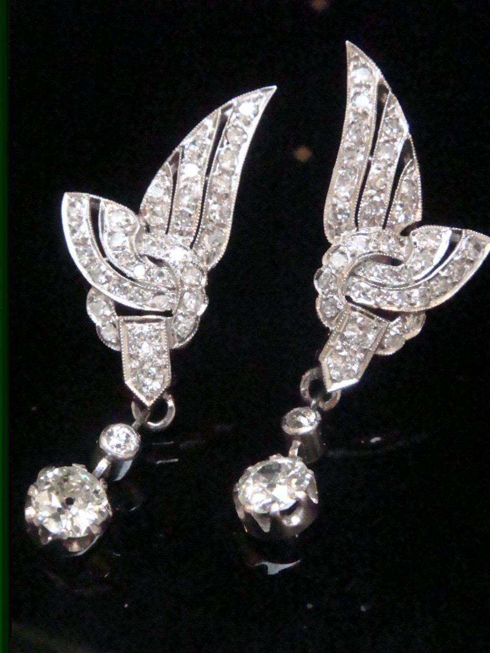 A pair of Art Deco 18ct white gold drop earrings stylised with geometric wing motifs set with diamonds and suspending articulated round old-cut diamond drops.