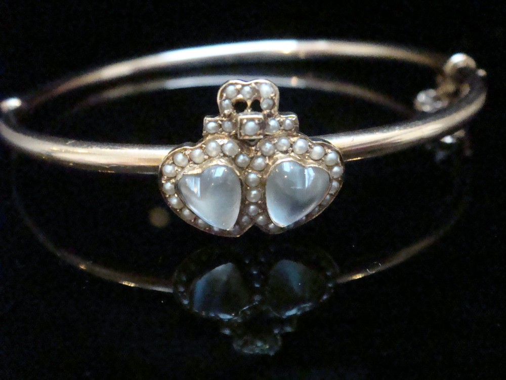 An antique 15ct gold hinged bangle featuring two heart-shaped cabochon moonstones framed by small seed pearls.