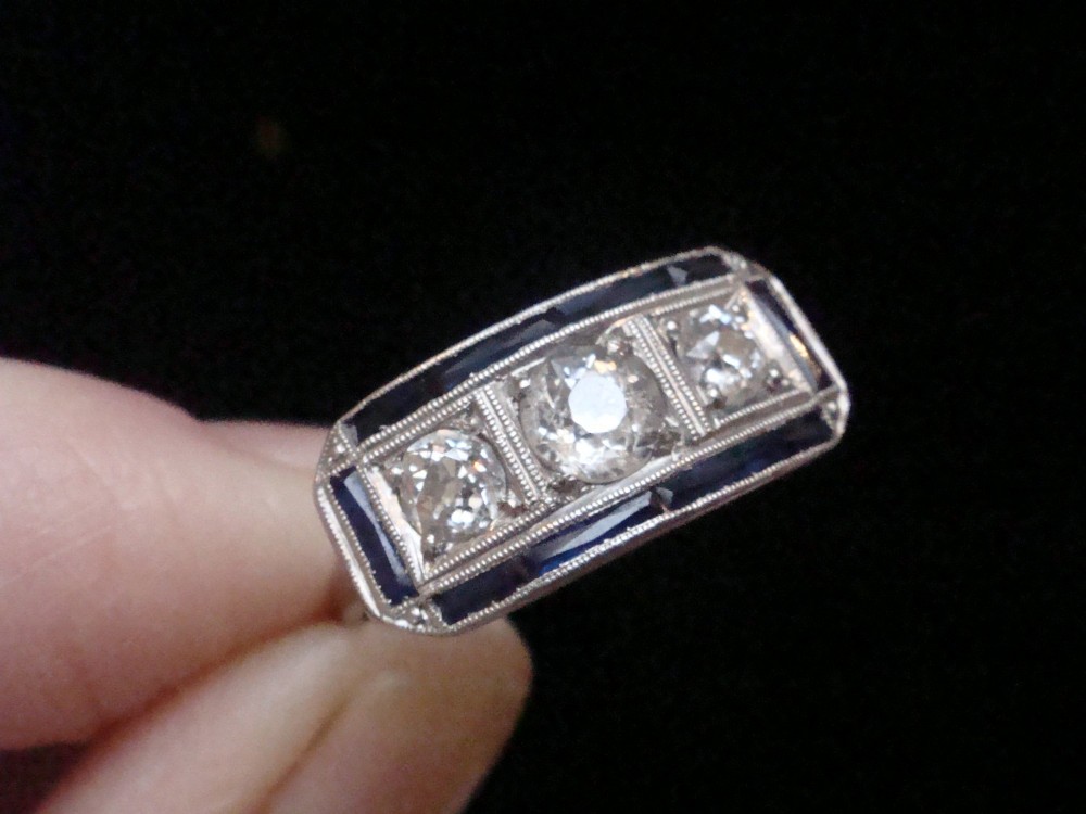 An Art Deco platinum ring featuring three central round diamonds flanked by dark blue calibre-cut sapphires in a rectangular millegrain setting. View IV