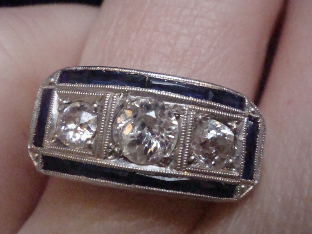 An Art Deco platinum ring featuring three central round diamonds flanked by dark blue calibre-cut sapphires in a rectangular millegrain setting. View V