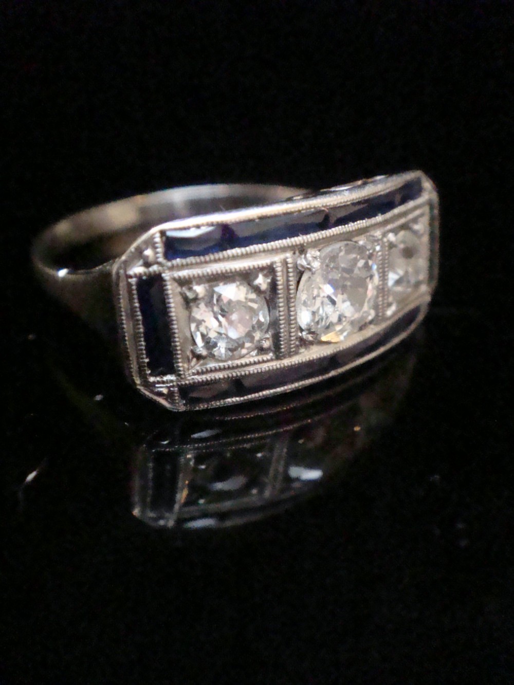 An Art Deco platinum ring featuring three central round diamonds flanked by dark blue calibre-cut sapphires in a rectangular millegrain setting. View VIII