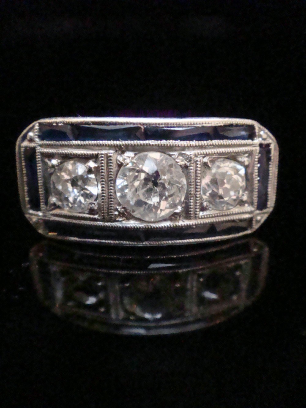 An Art Deco platinum ring featuring three central round diamonds flanked by dark blue calibre-cut sapphires in a rectangular millegrain setting. View IX