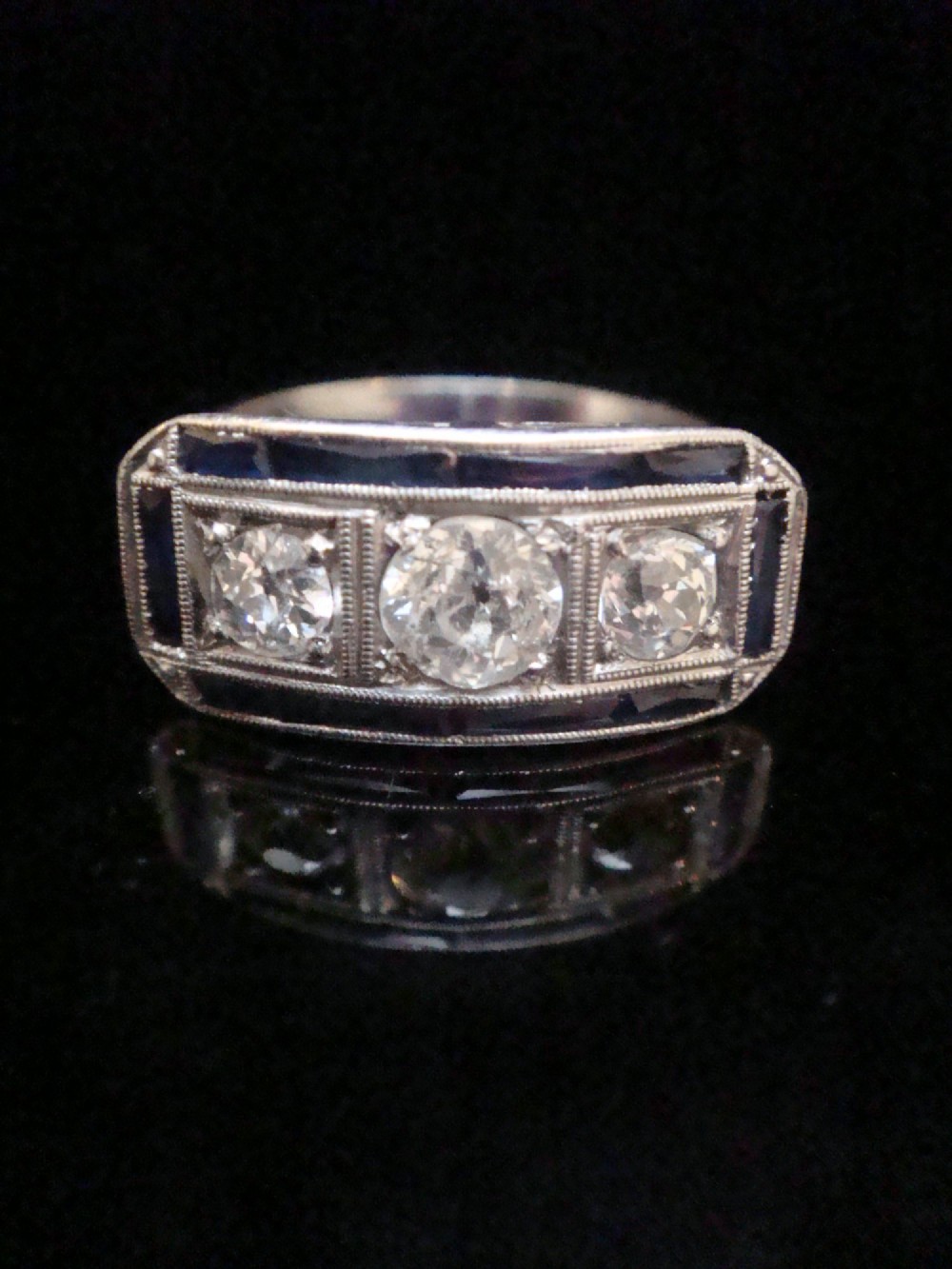An Art Deco platinum ring featuring three central round diamonds flanked by dark blue calibre-cut sapphires in a rectangular millegrain setting.