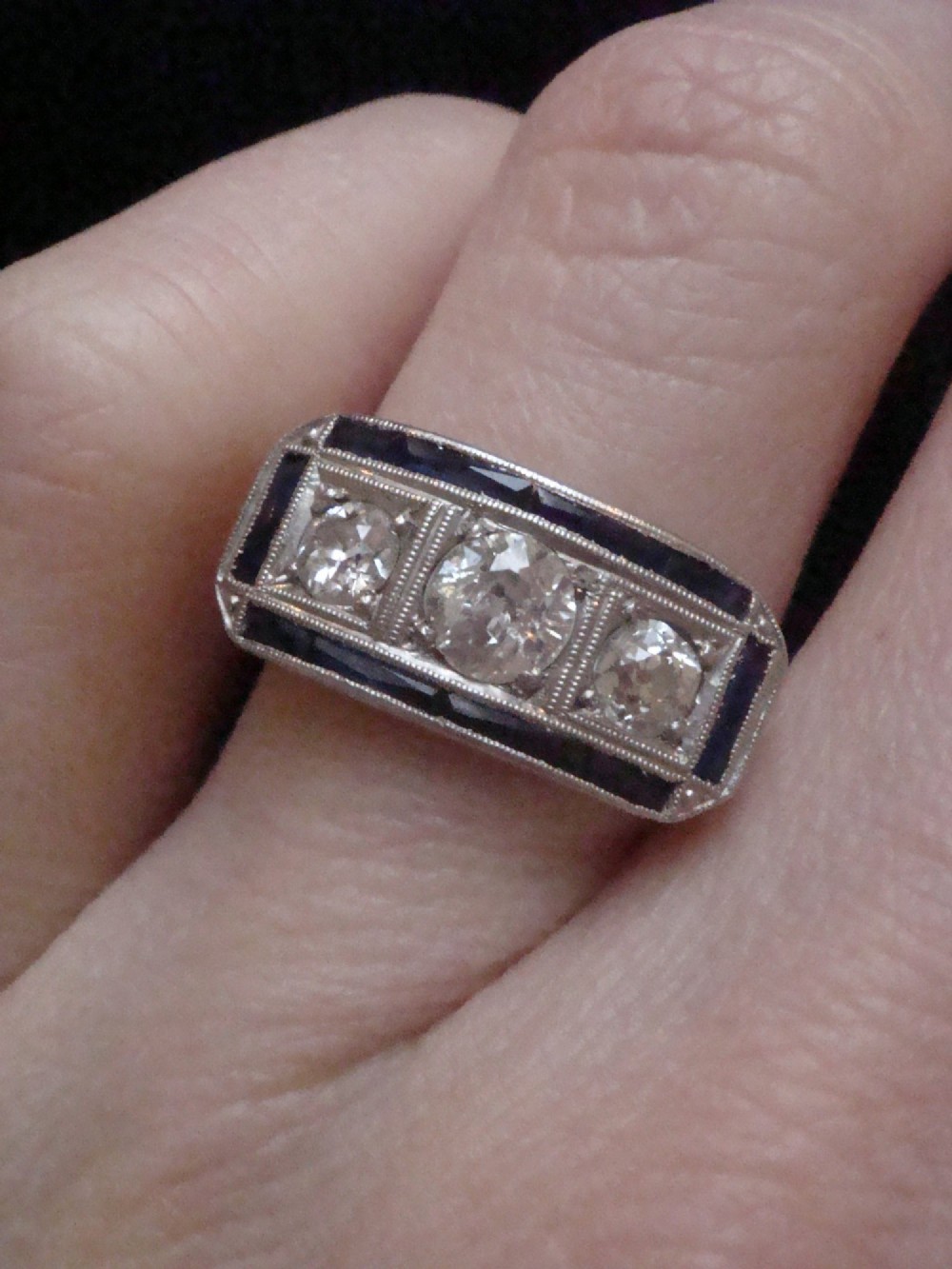 An Art Deco platinum ring featuring three central round diamonds flanked by dark blue calibre-cut sapphires in a rectangular millegrain setting. View VI