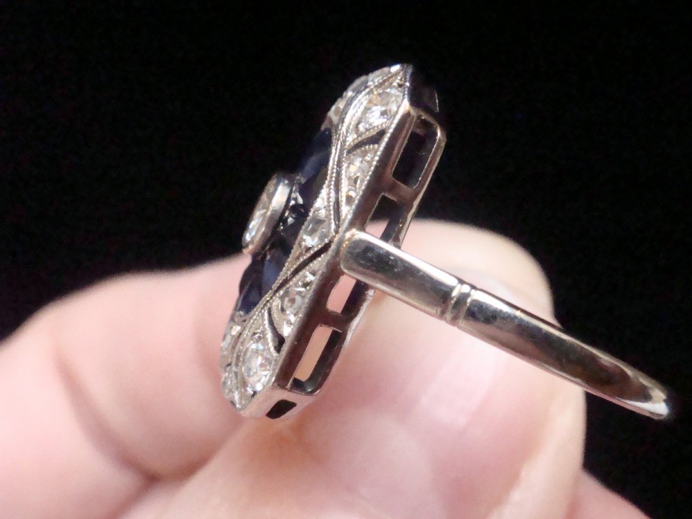 An Art Deco rectangular white gold ring featuring a central round diamond surrounded by calibre-cut blue sapphires and framed by smaller diamonds with detailed millegrain edging. View II
