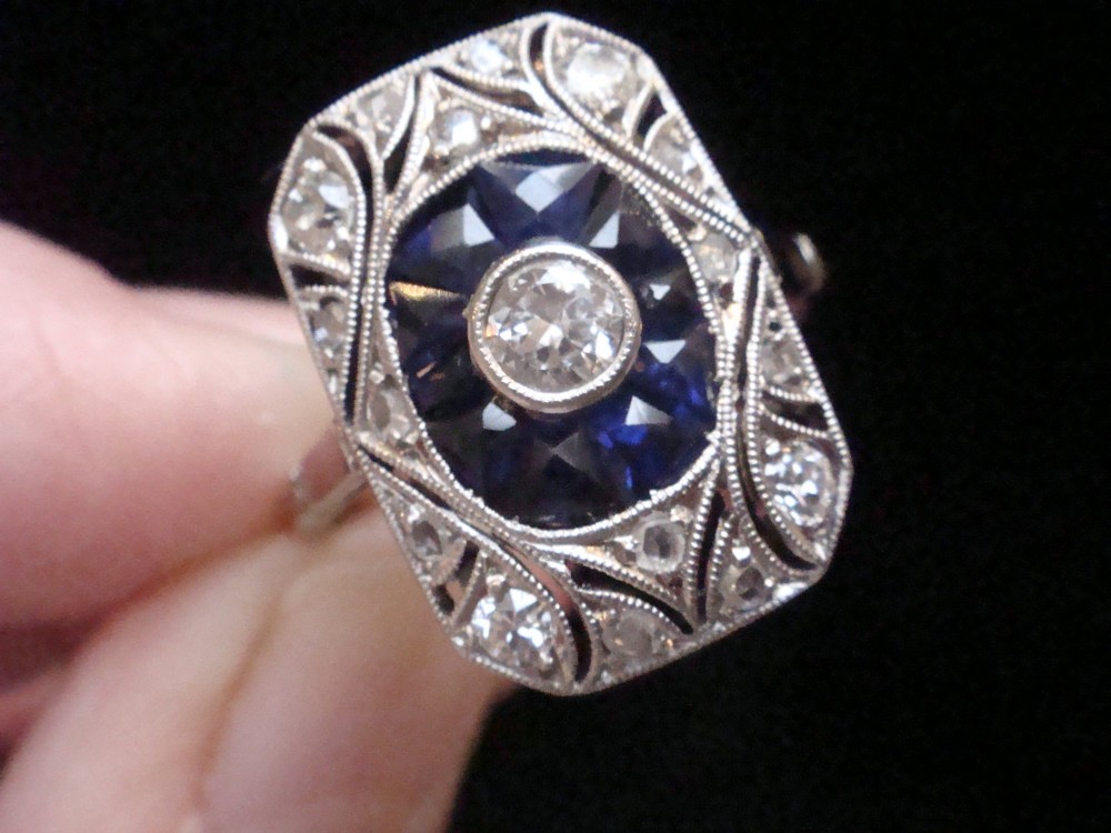 An Art Deco rectangular white gold ring featuring a central round diamond surrounded by calibre-cut blue sapphires and framed by smaller diamonds with detailed millegrain edging. View IV